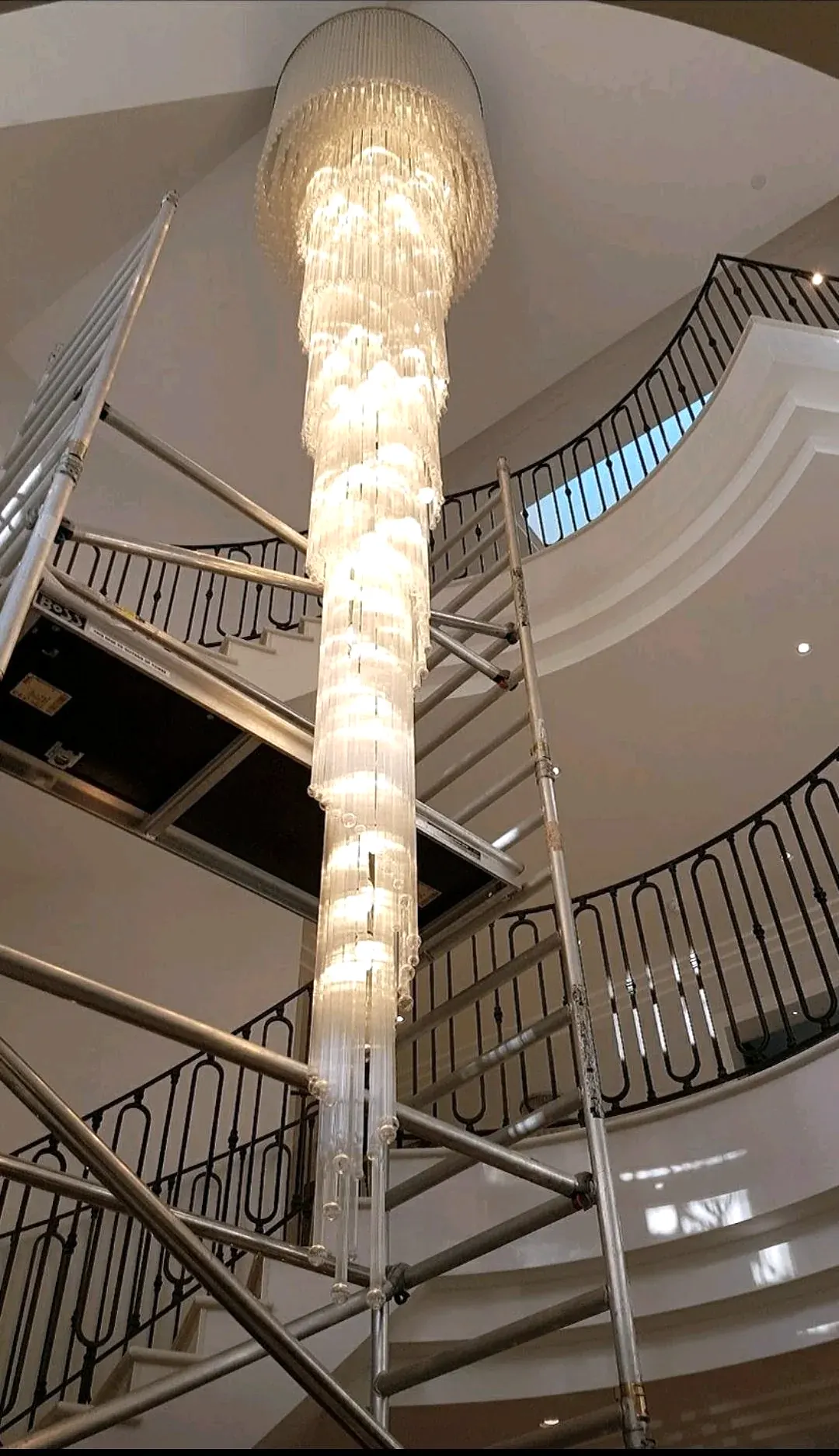 Tall crystal tube cascade chandelier in a curved stairwell, photographed from below with access scaffolding around it