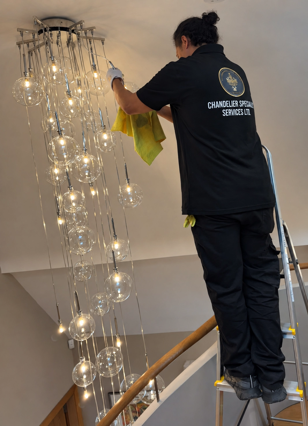 A specialist on a ladder cleaning a cascade of clear glass globe pendants above a staircase