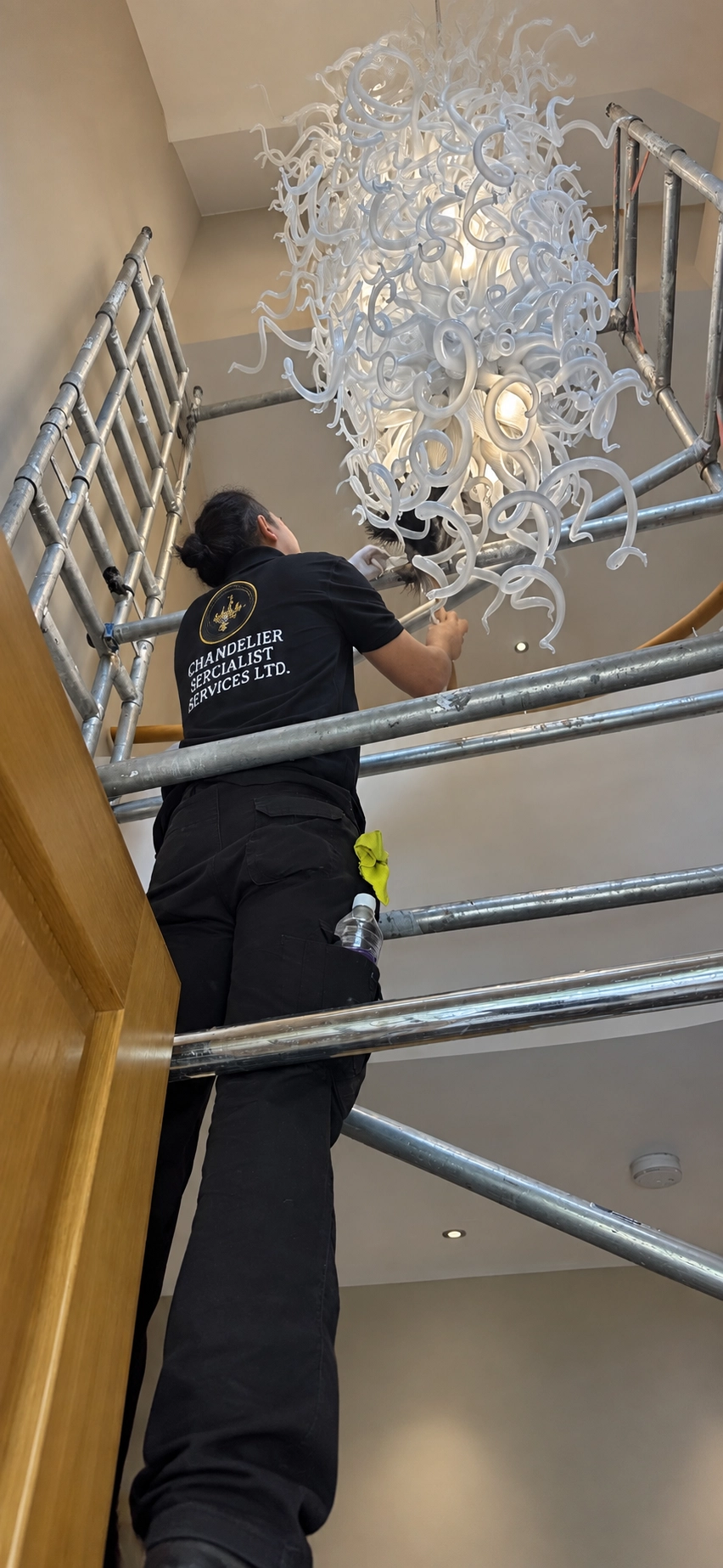 Chandelier specialist working from scaffolding beneath a white hand-blown glass swirl chandelier