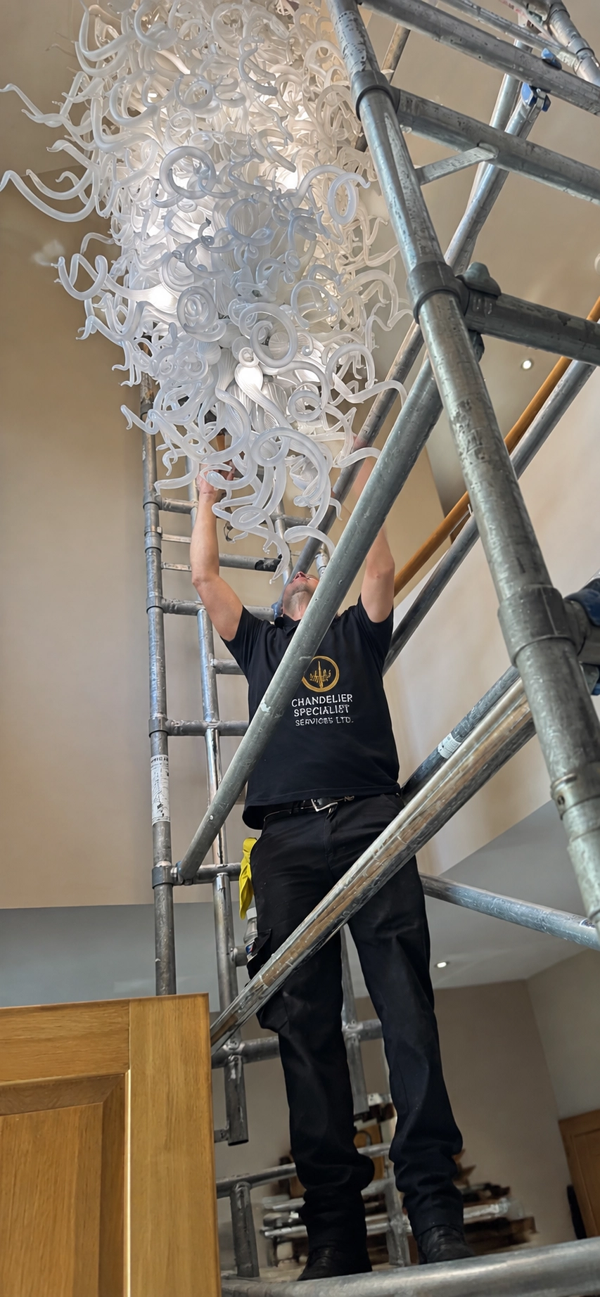 Chandelier specialist working from scaffolding on a white hand-blown glass swirl chandelier in a wider view